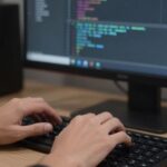 Developer typing code on a keyboard, monitor in background.
